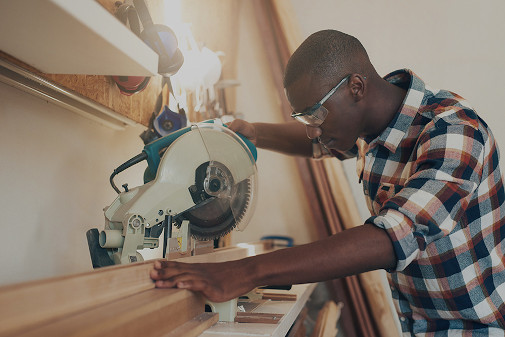 Young aritsan of African descent working with expertise in a woodworking workshop, handling a circular saw, and wearing safety goggles